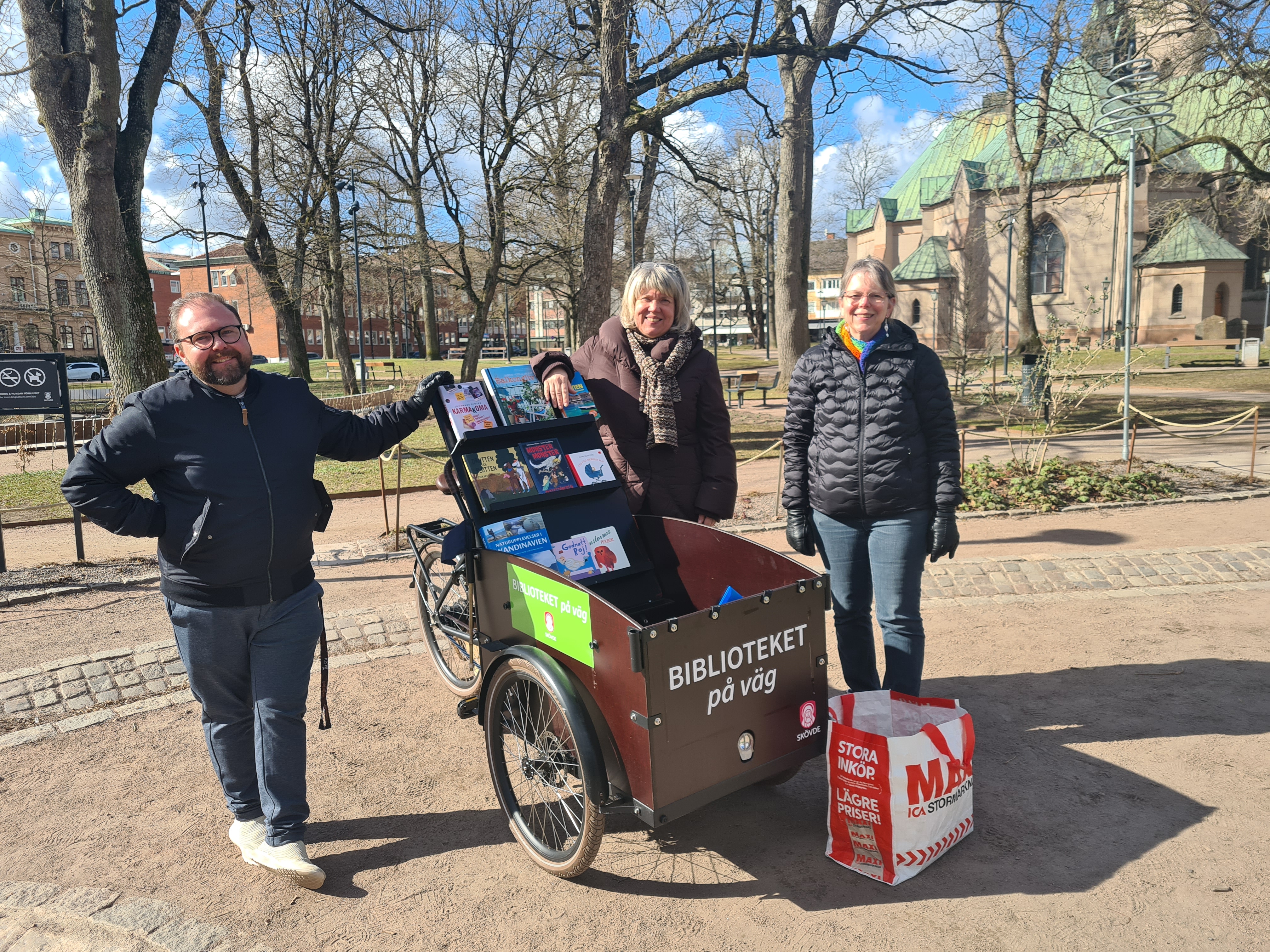 En man och två kvinnor som står vid en bokcykel fylld med böcker. På cykeln står det "Biblioteket på väg"